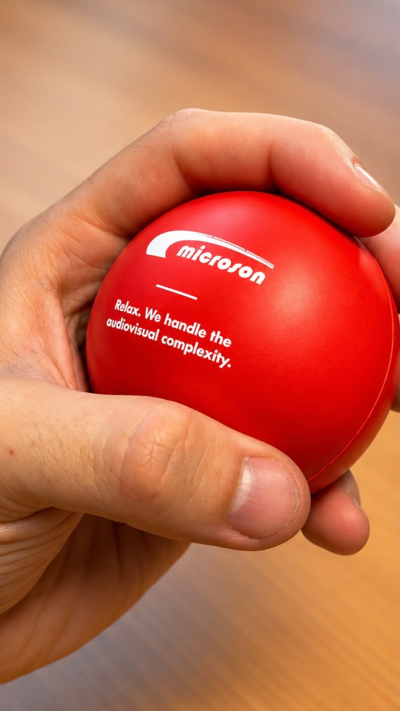Red Microson stress ball held in a hand, with the message “Relax. We handle the audiovisual complexity.”